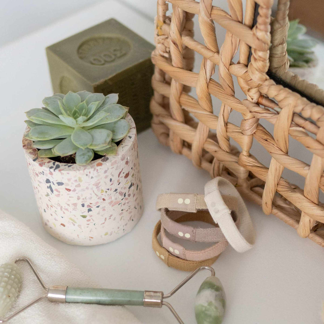 Plastic-free hair ties in neutral colors stacked on vanity surrounded by facial roller, potted plant, and mirror.