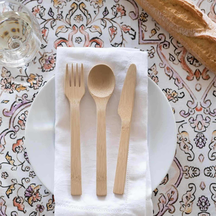 Bamboo utensil set, shown separately with fork, spoon, and knife atop white napkin on white plate, surrounded by print tablecloth, glass of white wine at top left, and baguette at top right.