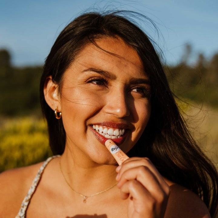 Woman applying lip balm in compostable tube,  standing outdoors.