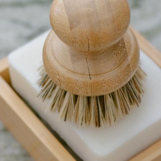 Plastic-free pot scrubber brush atop bamboo soap shelf and solid soap.
