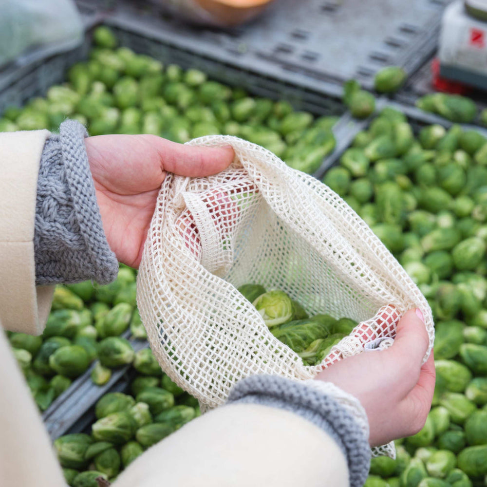 EcoBags plastic-free reusable produce bag, shown at market in person's hands with brussels sprouts in bag and below in gray crates.