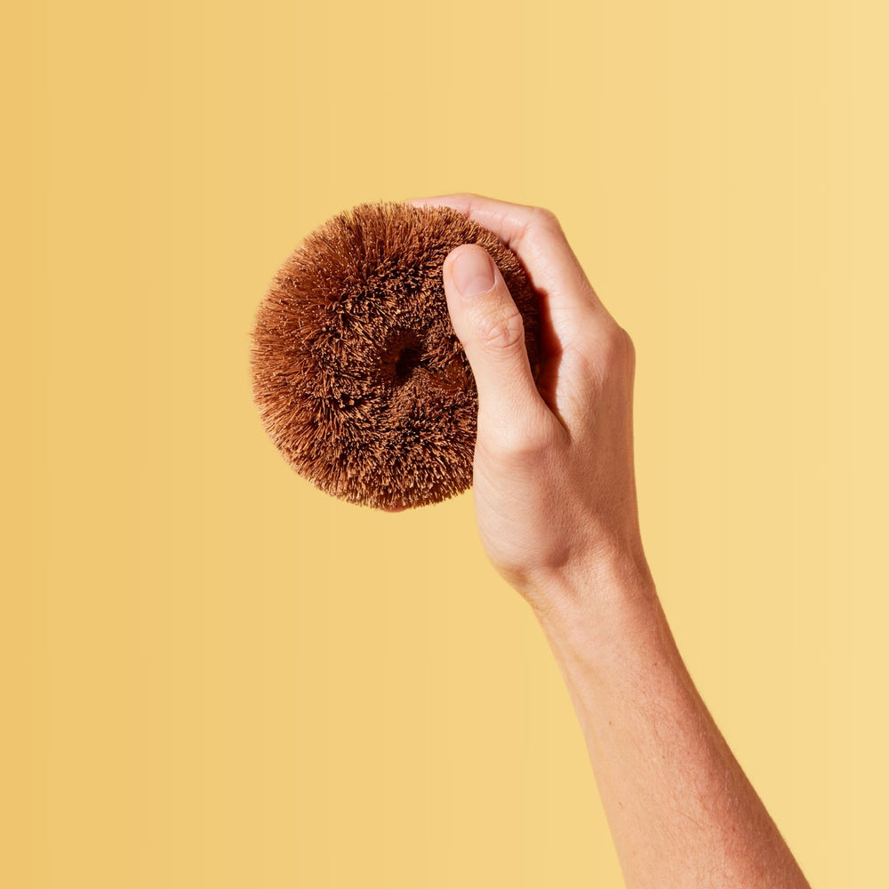 Coconut fiber scouring and scrubbing tool, shown in hand with yellow background.