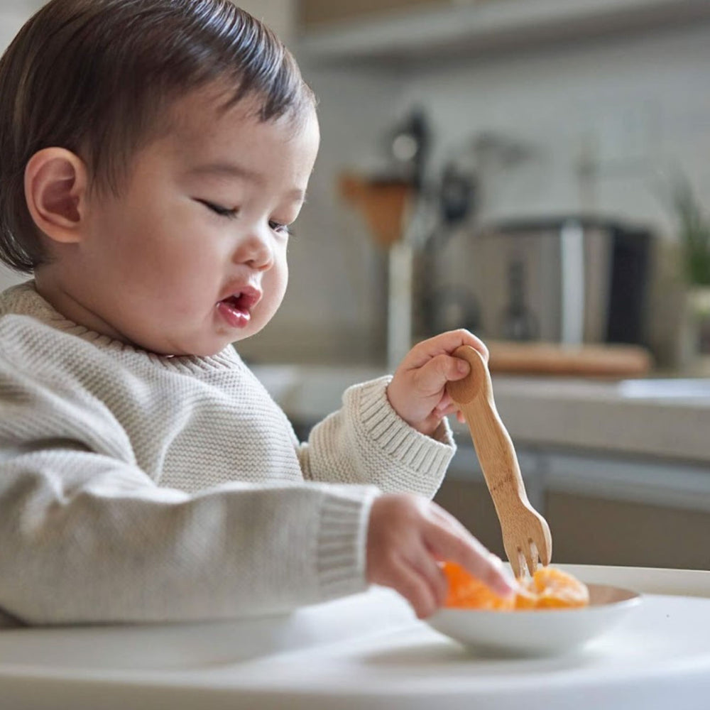 Kid's bamboo spork, shown with child eating orange sections from small white bowl and kitchen countertop in background.