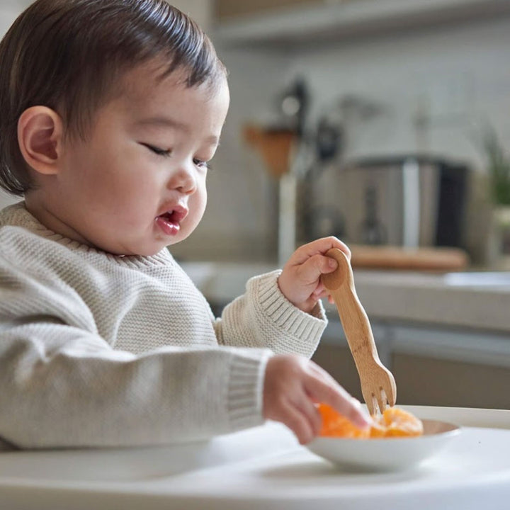 Kid's bamboo spork, shown with child eating orange sections from small white bowl and kitchen countertop in background.