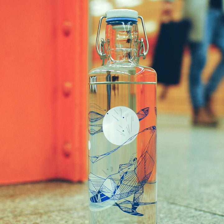 Soulbottles Souldiver Glass Water Bottle. 1.0 Liter Capacity, Plastic Free Bottle with Blue and White Whale Design. Set on floor next to orange I-beam with people in background.
