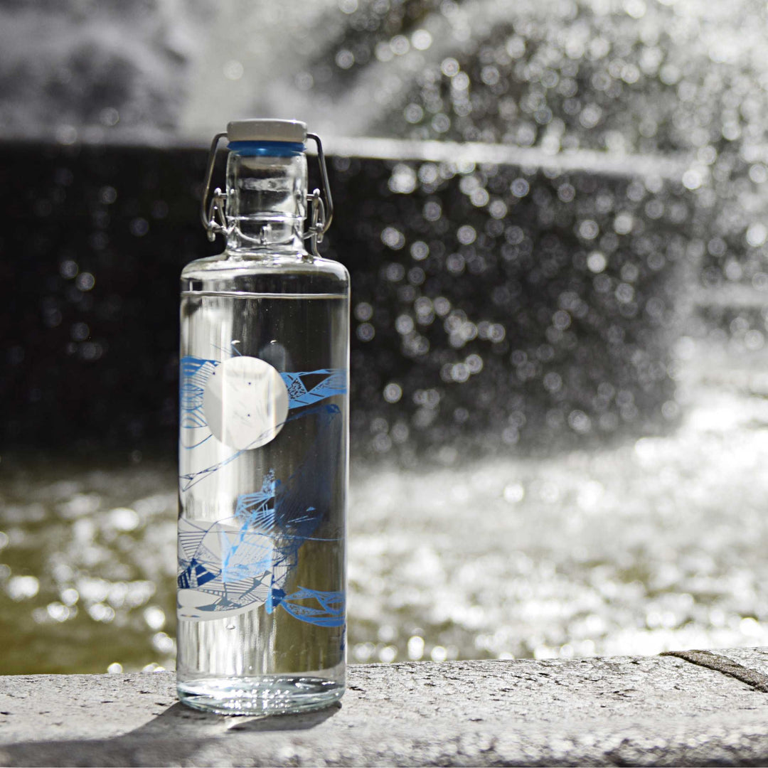 Soulbottles Souldiver Glass Water Bottle. 1.0 Liter Capacity, Plastic Free Bottle with Blue and White Whale Design. Set on edge of water fountain.