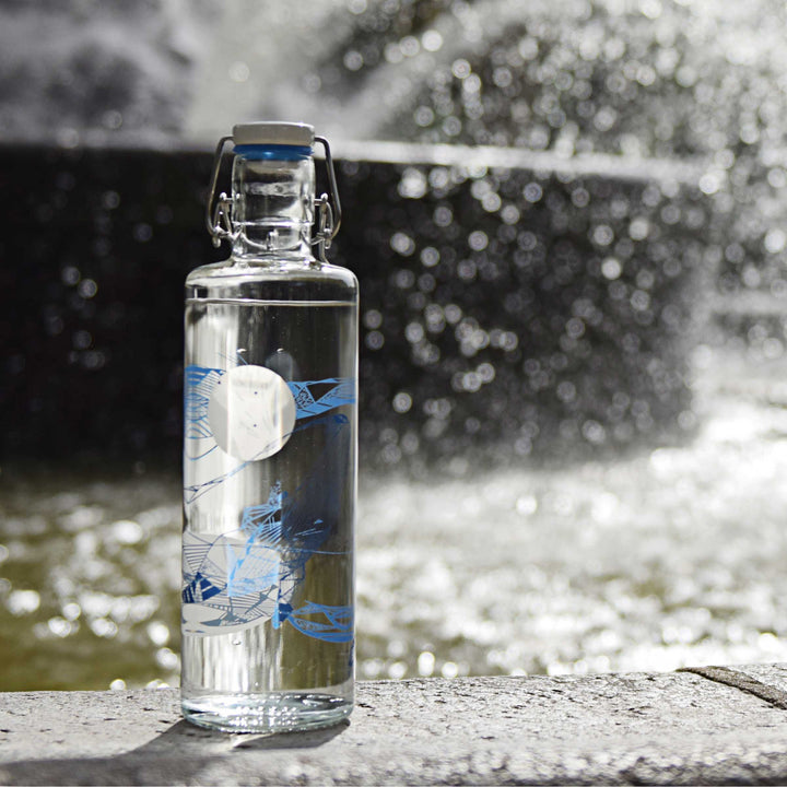 Soulbottles Souldiver Glass Water Bottle. 1.0 Liter Capacity, Plastic Free Bottle with Blue and White Whale Design. Set on edge of water fountain.