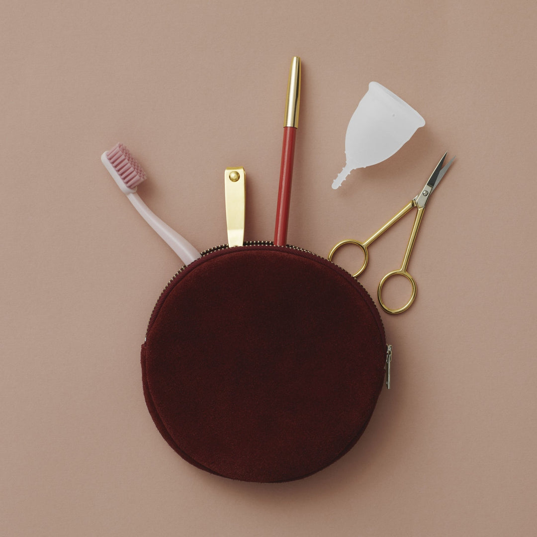 Menstrual cup on upper right, shown with round red makeup bag, toothbrush, nail clippers, makeup pencil, and small scissors for size reference.