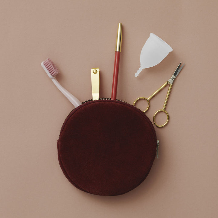 Menstrual cup on upper right, shown with round red makeup bag, toothbrush, nail clippers, makeup pencil, and small scissors for size reference.
