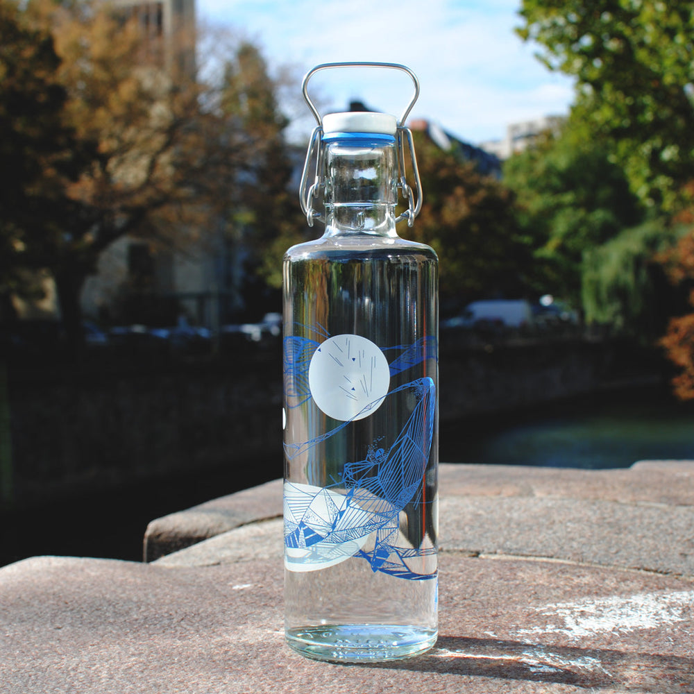 Soulbottles Souldiver Glass Water Bottle. 1.0 Liter Capacity, Plastic Free Bottle with Blue and White Whale Design. Set on Large Rock with Trees in Background.