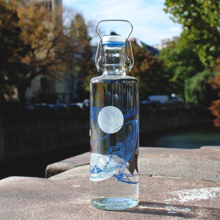 Soulbottles Souldiver Glass Water Bottle. 1.0 Liter Capacity, Plastic Free Bottle with Blue and White Whale Design. Set on Large Rock with Trees in Background.