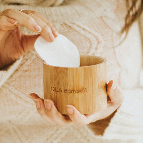 OLA Bamboo Eco Friendly Reusable Makeup Remover Pad Set With Container And Mesh Wash Bag. Container Held In Woman's Hand, While Removing A Round With Other Hand.